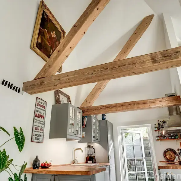 Trækonstruktion i køkken med synlige loftsbjælker over grå skabe og en træbordplade. Dekoreret med planter, maleri og skilt med teksten: "This kitchen is for dancing. Be Happy."