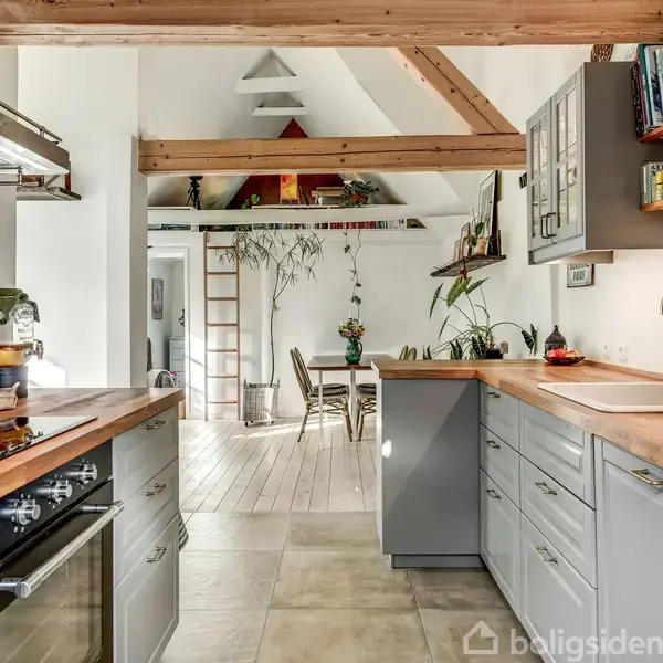 Køkken med træbordplader, grå skabe og indbyggede apparater. I baggrunden står et spisebord omgivet af planter. Store bjælker pryder loftet i et lyst rum med naturligt lysindfald.