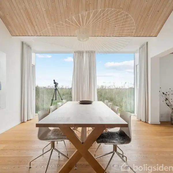 Et langt spisebord med stole står i et lyst rum med store vinduer. Et maleri hænger på væggen, og en vase samt et teleskop ses i baggrunden.