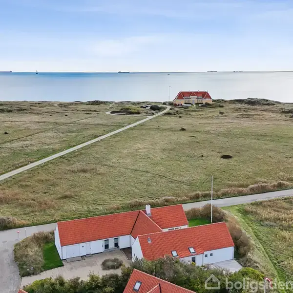 Et hvidt hus med rødt tag står stille, omgivet af en grøn eng. I baggrunden strækker vejen sig mod kysten og det lyseblå hav med flere skibe i horisonten.
