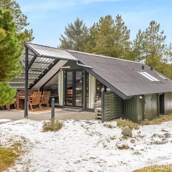 Sommerhus med skråt tag og store vinduespartier står på en snedækket grund omgivet af fyrretræer. En overdækket terrasse har et træbord og stole.