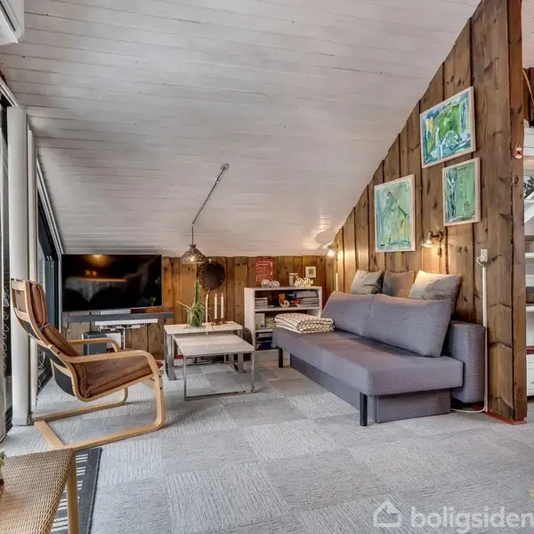 En stue med skråt loft indeholder en grå sofa, et tv på en reol, et lille bord og træpaneler. Danske flag og planter dekorerer rummet, som har store vinduer.
