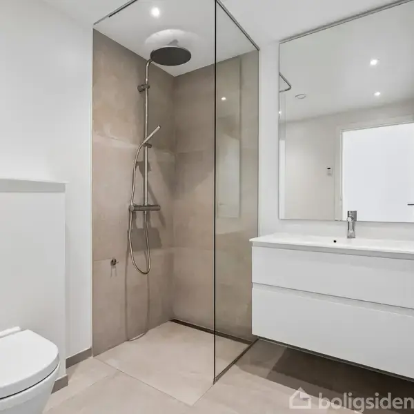 Et moderne badeværelse indeholder et væghængt toilet, en brusekabine med klart glas og regnbruser, samt en hvid håndvask med skab og stort spejl. Rummet er minimalistisk med lys belysning.