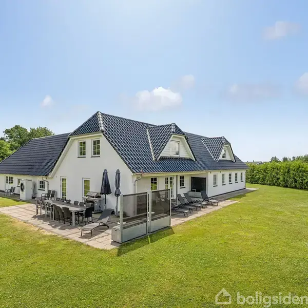 Hvidt hus med sort tag står stille på en græsplæne. Ved siden af er en terrasse med havemøbler og en parasol. Omgivet af træer og hække under en blå himmel.