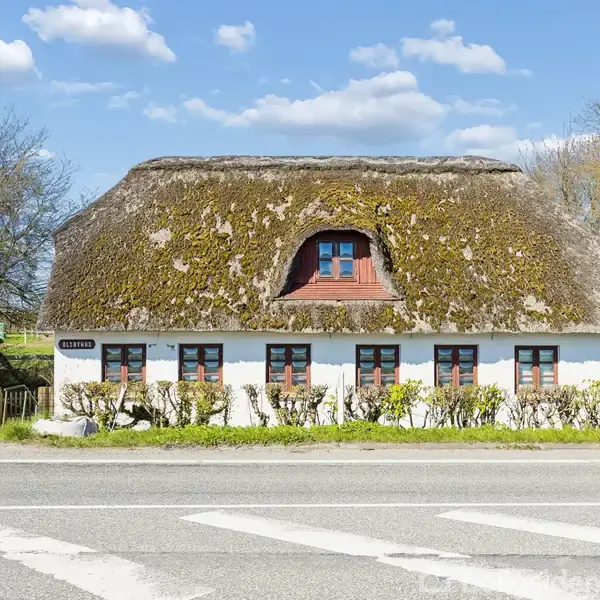 Hus med stråtag og hvide vægge står langs en vej, omgivet af træer. Skilt ved indgang viser teksten "Klitrose". Skyer og blå himmel i baggrunden.