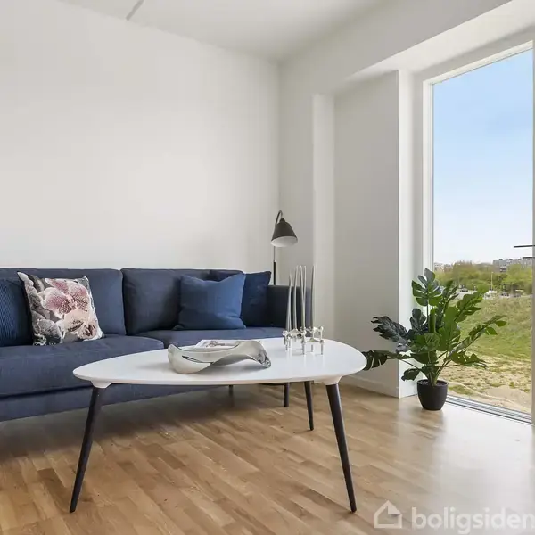 En blå sofa står ved siden af et sofabord med lysestager i en lys stue. Store vinduer viser udsigt til grønne områder. En gulvplante står ved døren.