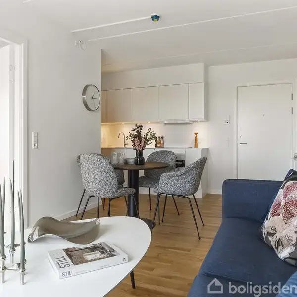 En stue med en blå sofa og pyntepuder, et rundt spisebord med stole, lyse køkkenskabe i baggrunden, og et hvidt sofabord med en kandelaber og en bog.