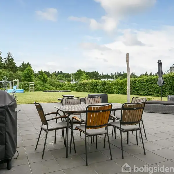 En terrasse med havemøbler og en grill placeret på gråt flisegulv. I baggrunden ses en grøn have med et legeområde og skov. Himlen er let skyet.