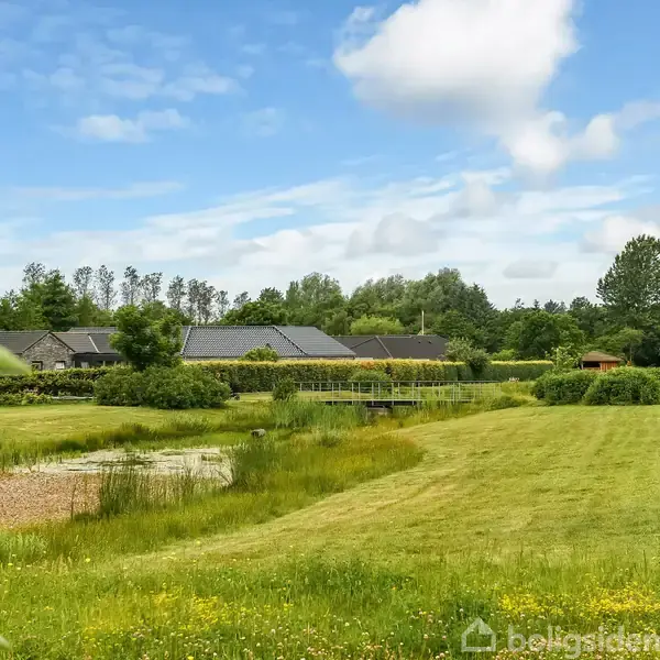 Græsplæne strækker sig mod række af huse i rolige omgivelser med blå himmel og enkelte skyer. En sø er placeret foran husene, omgivet af buske og træer.