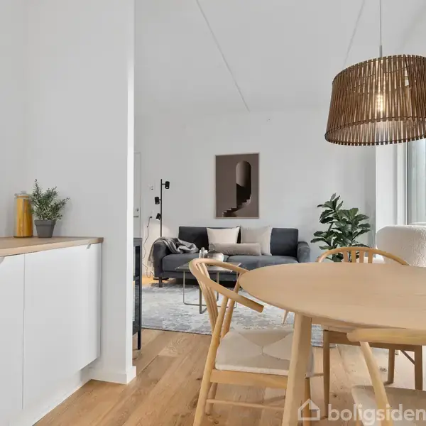 Et lyst køkkenalrum med et rundt spisebord og træstole; over bordet hænger en stor lampeskærm. I baggrunden ses en stue med en grå sofa og grønne planter.