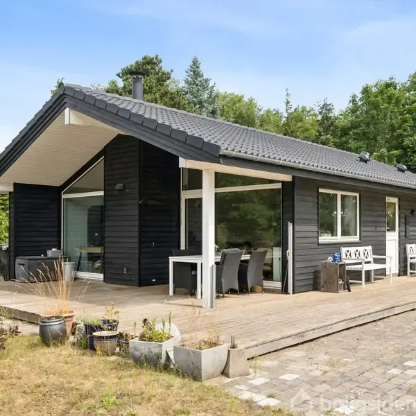 Et sort sommerhus med store vinduer står på en træterrasse omgivet af skov. Hvide havemøbler og blomsterkrukker er placeret udenfor, skaber en hyggelig atmosfære.