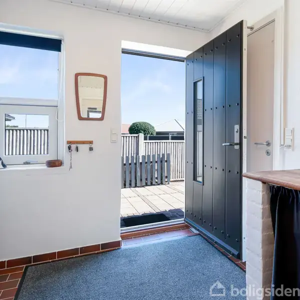 Døren står åben i en entré med udsigt til en terrasse. Væggene er lyse, et vindue og et spejl hænger ved siden af vasken. Skabe og skuffe er til venstre.