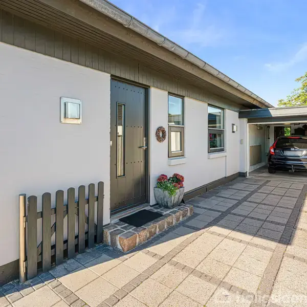 En carport med en sort bil holder til venstre for en moderne, hvid facade med flere vinduer. En lille træport og blomsterdekoration ses nær hoveddøren, omgivet af fliser.