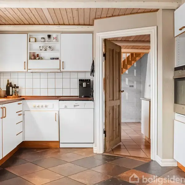 Køkken med hvide skabe og trækanter er udstyret med ovn, opvaskemaskine og kaffemaskine. Der hænger lamper over arbejdsbordet, og en åben dør fører til en trappeopgang.