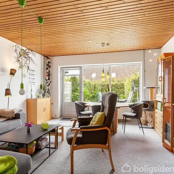 En stue med en grå hjørnesofa og lænestol ved et lavt sofabord. Store vinduer giver udsigt til have. Bogreoler og grønne pyntegenstande står rundt i rummet.