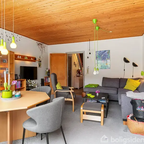 En stue indeholder en grå sofa med farverige puder, et sofabord, og en rund spisebord med stole. Omgivet af vægge dekoreret med kunst, under et træloft.