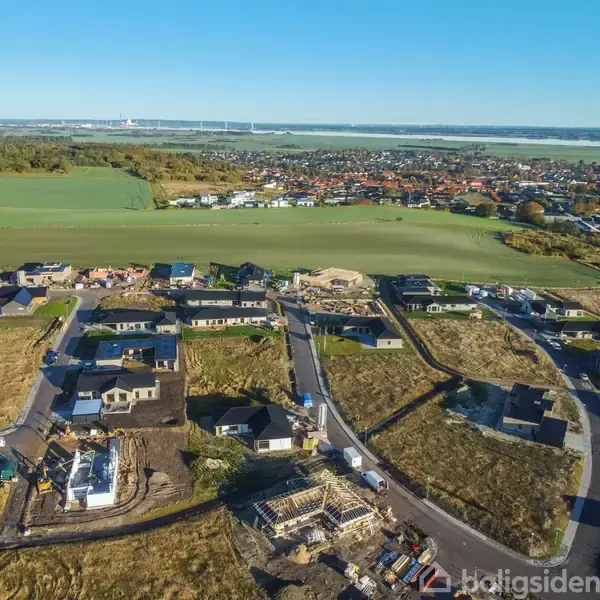 Byggeplads med nye huse under opførelse omgivet af marker og en by i baggrunden. Veje snor sig gennem området, mens maskiner og materialer ses spredt rundt omkring.