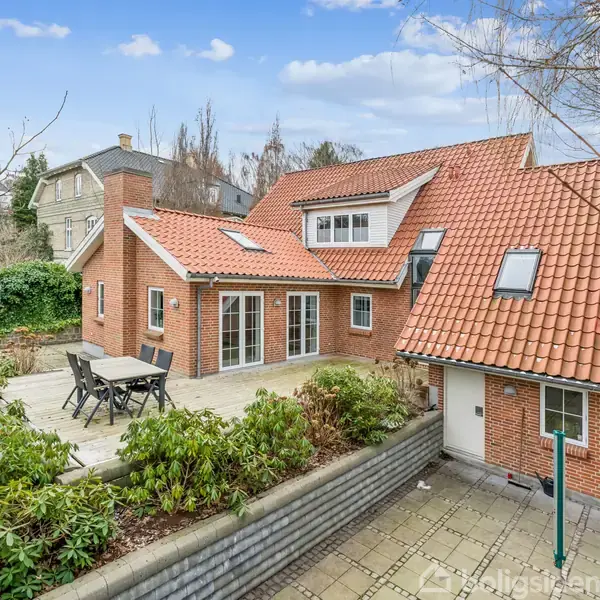 Rødt murstenshus med rødt tegltag, havebord og stole på terrasse. Haven har grønne buske og stenbelagte stier. Omgivet af mere beplantning og nabohuse under en blå himmel.