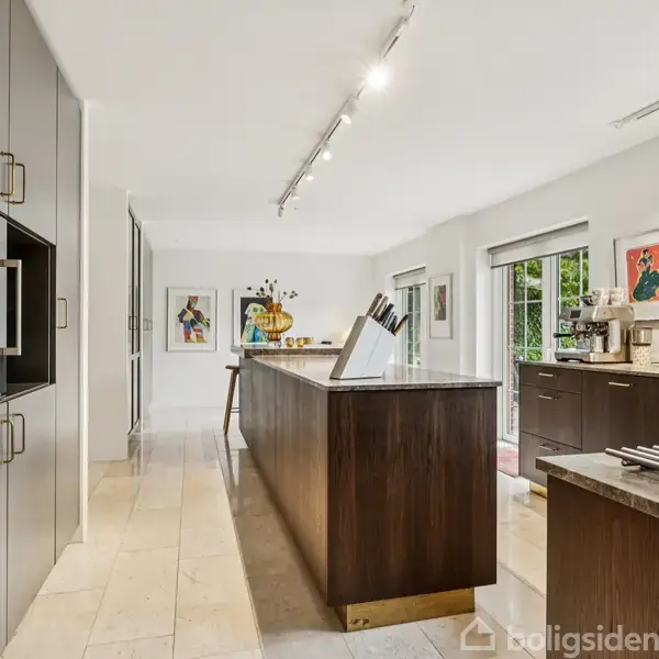 Et moderne køkken med en stor, mørk træø i midten, flankeret af grå skabe og sorte apparater. Kunstmalerier hænger på væggen, og store vinduer giver udsigt til grønne træer.