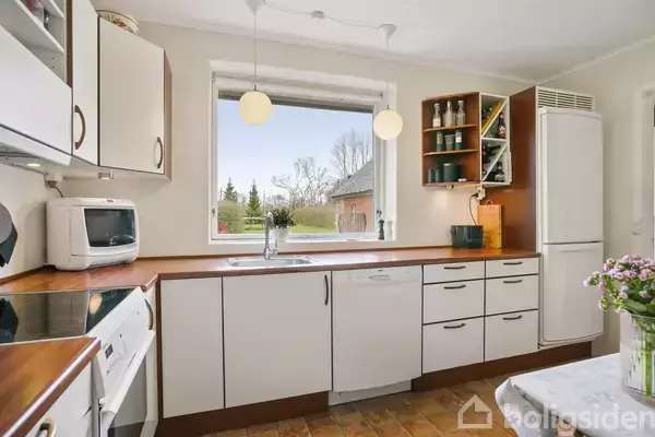 Et køkken med hvide skabe og træbordplader har opvaskemaskine og mikrobølgeovn under et stort vindue. Udsigt til have med grønt græs og træer udenfor. En vase med blomster står på et bord.