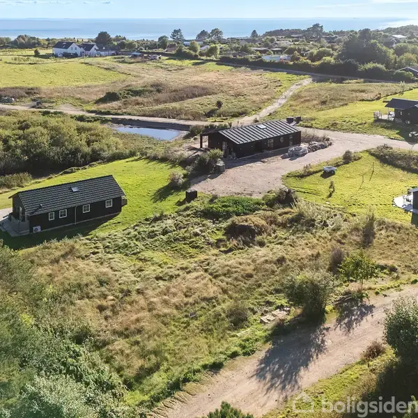 Flere huse med sorte tage ligger spredt i et bakket, grønt landskab nær havet. Sollys belyser området, der også inkluderer en grusvej og frodig vegetation.