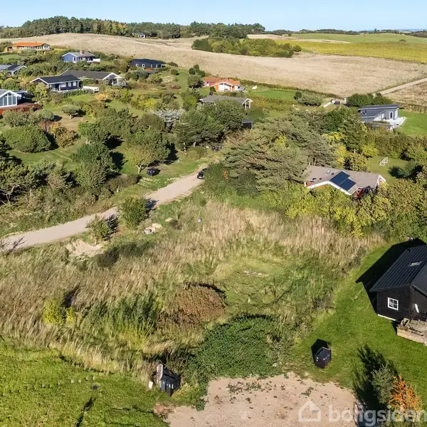 Sommerhuse strækker sig over et bakket landskab dækket af græs og træer. Nogle huse har solpaneler på taget, omgivet af haver og marker. En vej snor sig mellem husene.