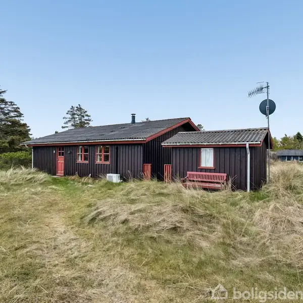 Et sort træhus med rødt tag og detaljer står midt i en græsmark. En antenne er på taget. Omkring huset er der højt græs og nogle træer i baggrunden.
