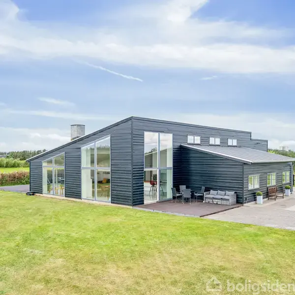 Sort, moderne enfamiliehus med store vinduespartier står på en velplejet græsplæne. Terrasse med havemøbler. Lyst, klart vejr omkranser det åbne landskab i baggrunden.