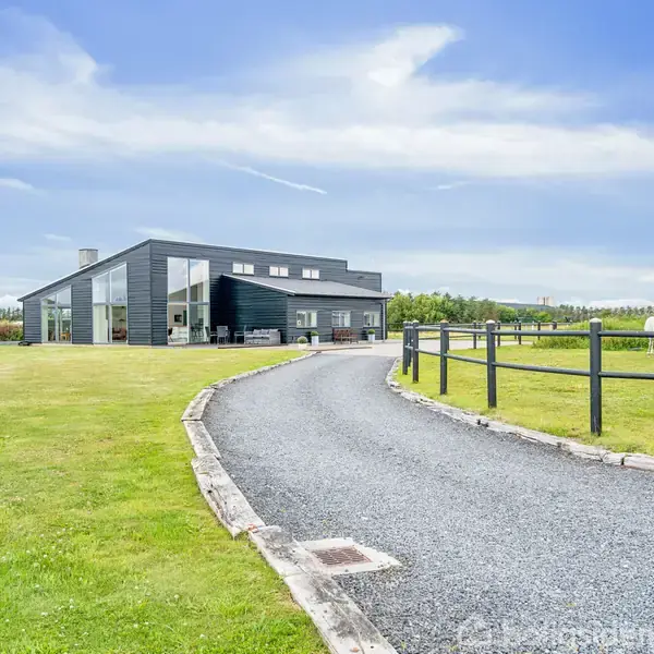 Et moderne, sort hus står ved siden af en snoet indkørsel. To heste græsser i en indhegning på en grøn mark under en blå himmel med lette skyer.