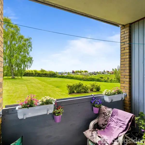 En balkon viser flere blomster i blomsterkasser, der er fastgjort til rækværket, hvor en stol med tæppe står. I baggrunden ses en græsplæne og et træ med blå himmel.