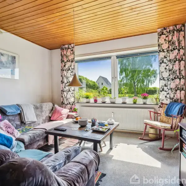 Stue med stor grå sofa og sofabord, dekoreret med puder og tæpper. Vindue med blomstrede gardiner viser udsigt til haver. Lyse træmøbler og akvarium ved siden. Maleri på væggen.