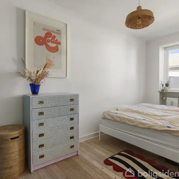 Et soveværelse med en seng til højre, en lyseblå skænk med seks skuffer til venstre. På skænken står en vase med tørrede blomster og en plakat med teksten "Lolita".