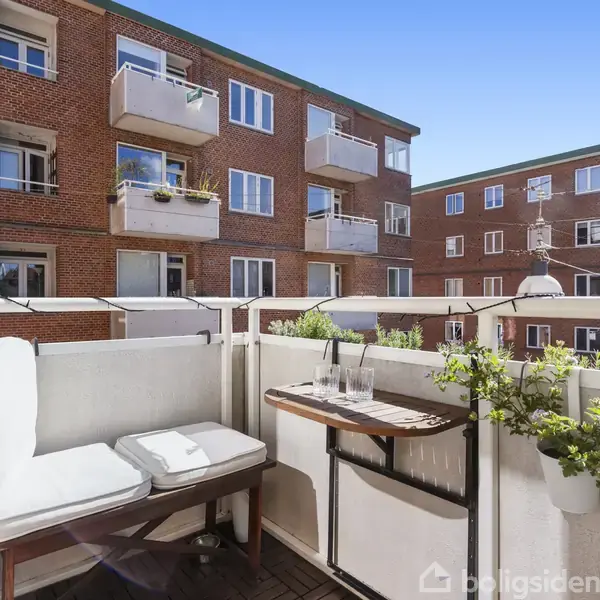 Balkon med hvid bænk og pude, bord med glas og kande, samt planter. Omgivet af murstensbygninger med altaner under en klar blå himmel.