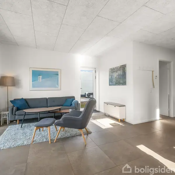 Stue med grå sofa og lænestol ved et sofabord, placeret på et lyst tæppe. Sollys strømmer ind gennem store vinduer, med en plante til venstre og lys kunst på væggen.