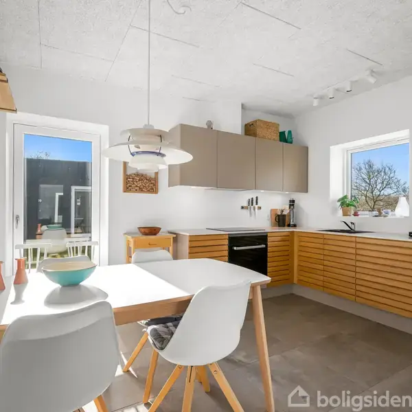 Et moderne køkken med et hvidt bord og to stole. Bordet har skåle, og pladsen er oplyst af en loftlampe. Store vinduer giver udsigt til en terrasse og træer udenfor.