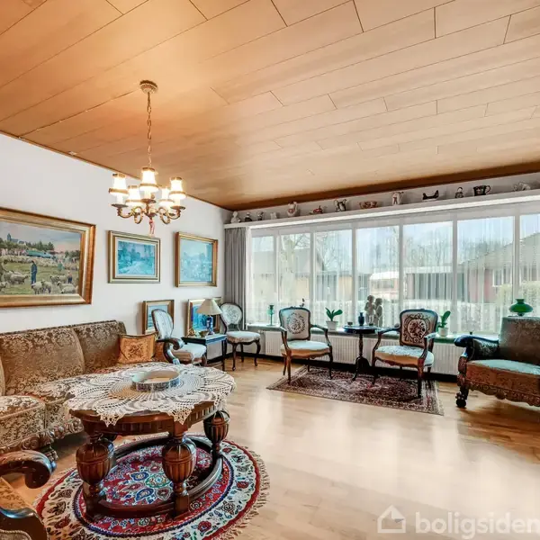 Stue med brun sofa og antikt sofabord, dekoreret med malerier på væggen. Store vinduer med udsigt til haven og gulvlaminat i lyst træ. En lysekrone hænger fra loftet.