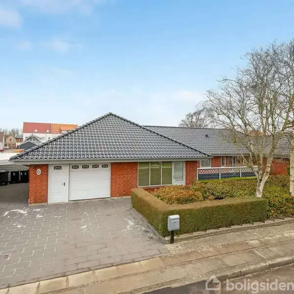 Huset er et rødt murstenshus med sort tegltag, to garageporte, og en flisebelagt indkørsel. Det er omgivet af en hæk og træer i en rolig boligkvarterskontekst.