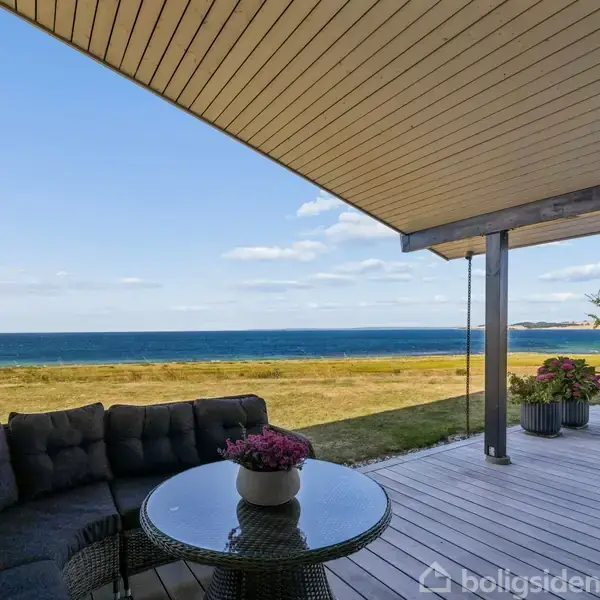 Terrassemøbler står på en træterrasse med udsigt over havet. En stor sofa med et lille bord og blomster er placeret under et halvtag, med græsplæne og kystlinje i baggrunden.