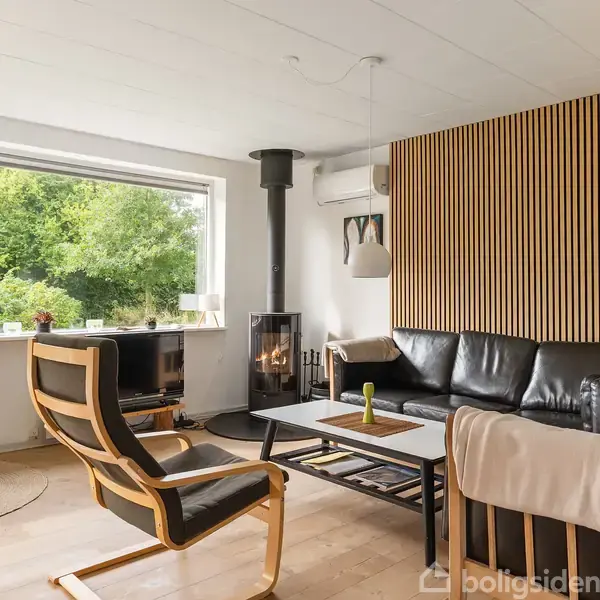 Stue med sorte lædersofaer og to træstole omkring et sofabord. En brændeovn tilføjer hygge. Store vinduer viser udsigt til grøn have. Trævægge og lamper skaber en moderne atmosfære.