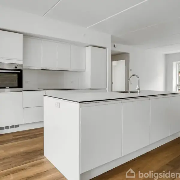 Køkken med hvidt interiør dominerer, mens en stor køkkenø indeholder en indbygget vask. I baggrunden ses et lyst rum med trægulve og adgang til en balkon.