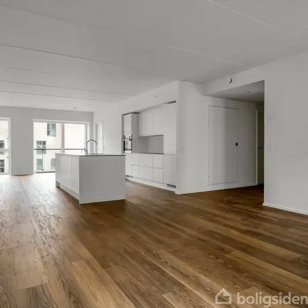 Et lyst, åbent køkkenalrum med trægulv har et køkkenø centralt. Store vinduer i baggrunden viser udsigt til en moderne bygning udenfor. Minimalistisk og indbydende atmosfære.