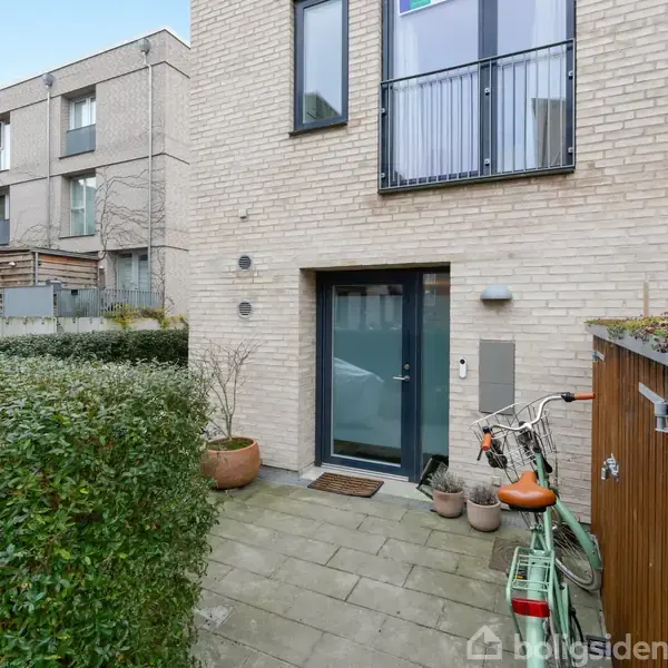 En moderne boligindgang med en glasdør omgivet af mursten. En grøn cykel står parkeret ved siden af en træskraldespand. Små potteplanter dekorerer fortovet i en urban boligområde.
