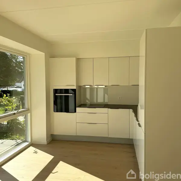 Et moderne hvidt køkken med indbyggede skabe og ovn placeret ved et stort vindue, som giver naturligt lysindfald. Udsigt til grønne planter udenfor.