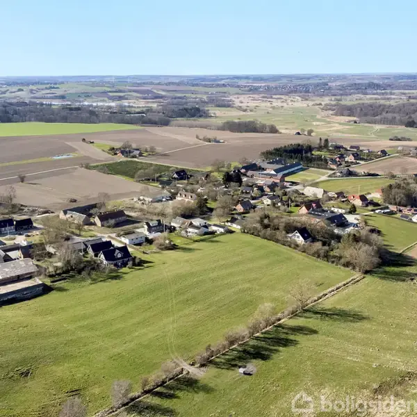Landsby omgivet af åbne marker og enge set ovenfra. Huse og gårde ligger spredt langs vejnettet. Langstrakte marker strækker sig i alle retninger, under en klar blå himmel.