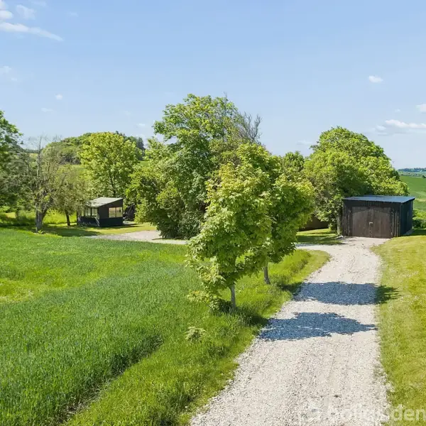 En grusvej snor sig gennem grønt landskab med spredte træer og to små, mørke bygninger omkranset af marker og en klar blå himmel.