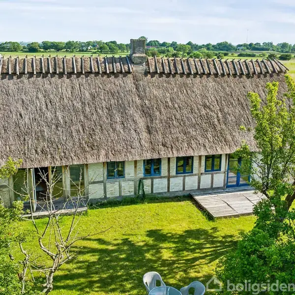 En stråtækt bindingsværkshus med skorsten står stille i en grøn have omkranset af træer. En terrasse er synlig, og landskabet strækker sig mod en horisont under en blå himmel.