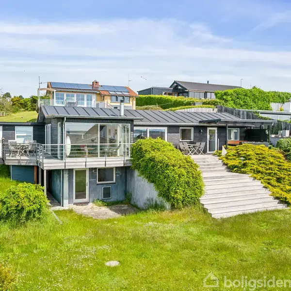 Et moderne hus med store glaspartier og balkon står på en grøn græsplæne med trapper. Omgivet af buske og flere huse i baggrunden under en blå himmel.