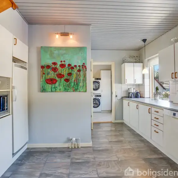 Et moderne køkken med hvide skabe og en lang bordplade. En farverig maleri af blomster hænger på væggen. I baggrunden ses en vaskesektion med vaskemaskine og tørretumbler.