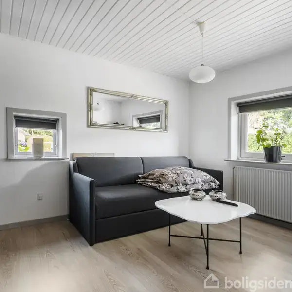 En grå sofa står i en lille stue med et sofabord foran. Rummet har et træloft, lyse trægulve, et spejl over sofaen og et vindue med en plante.