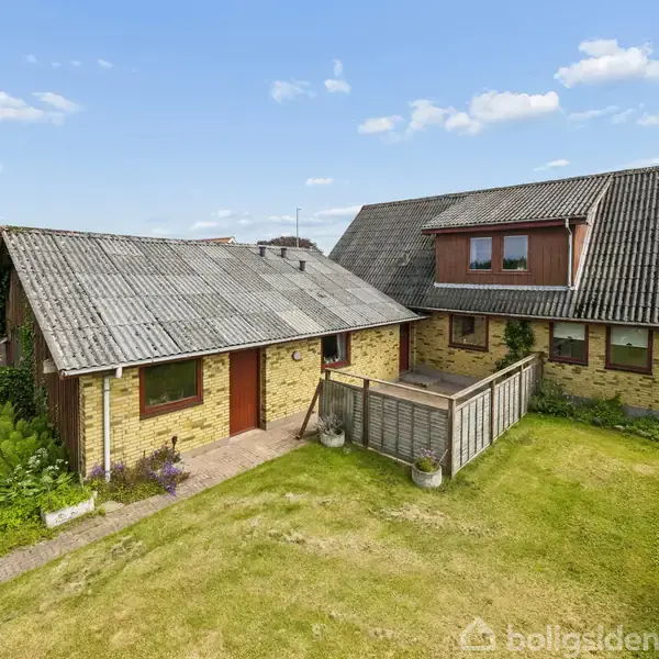 Et murstenshus med skråtag står stille i en grøn have. Ved siden af er der en lille træterrasse. Omkring huset er der plæner og buske. Himmel med lette skyer.